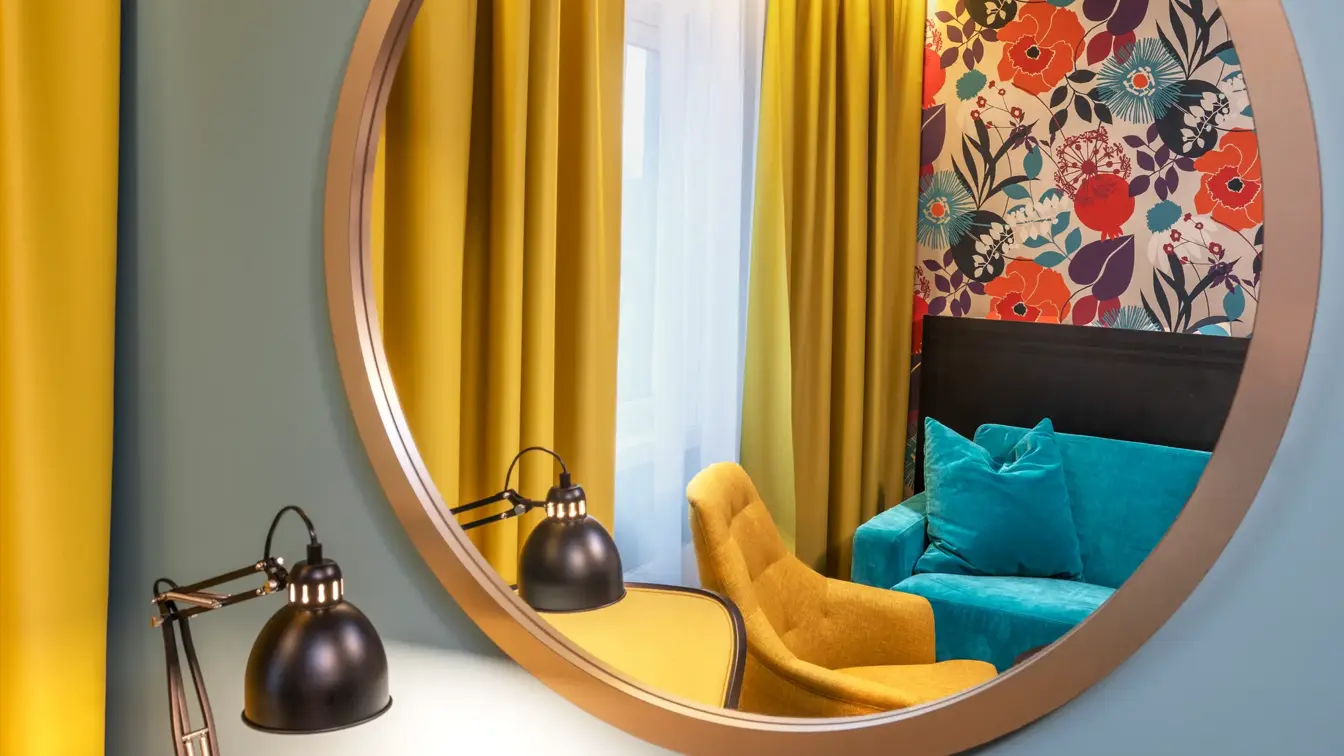 Svart skrivebord med en svart skrivesbordslampe, gul gardin, blå vegg med et rundt stort speil hengende på veggen. 