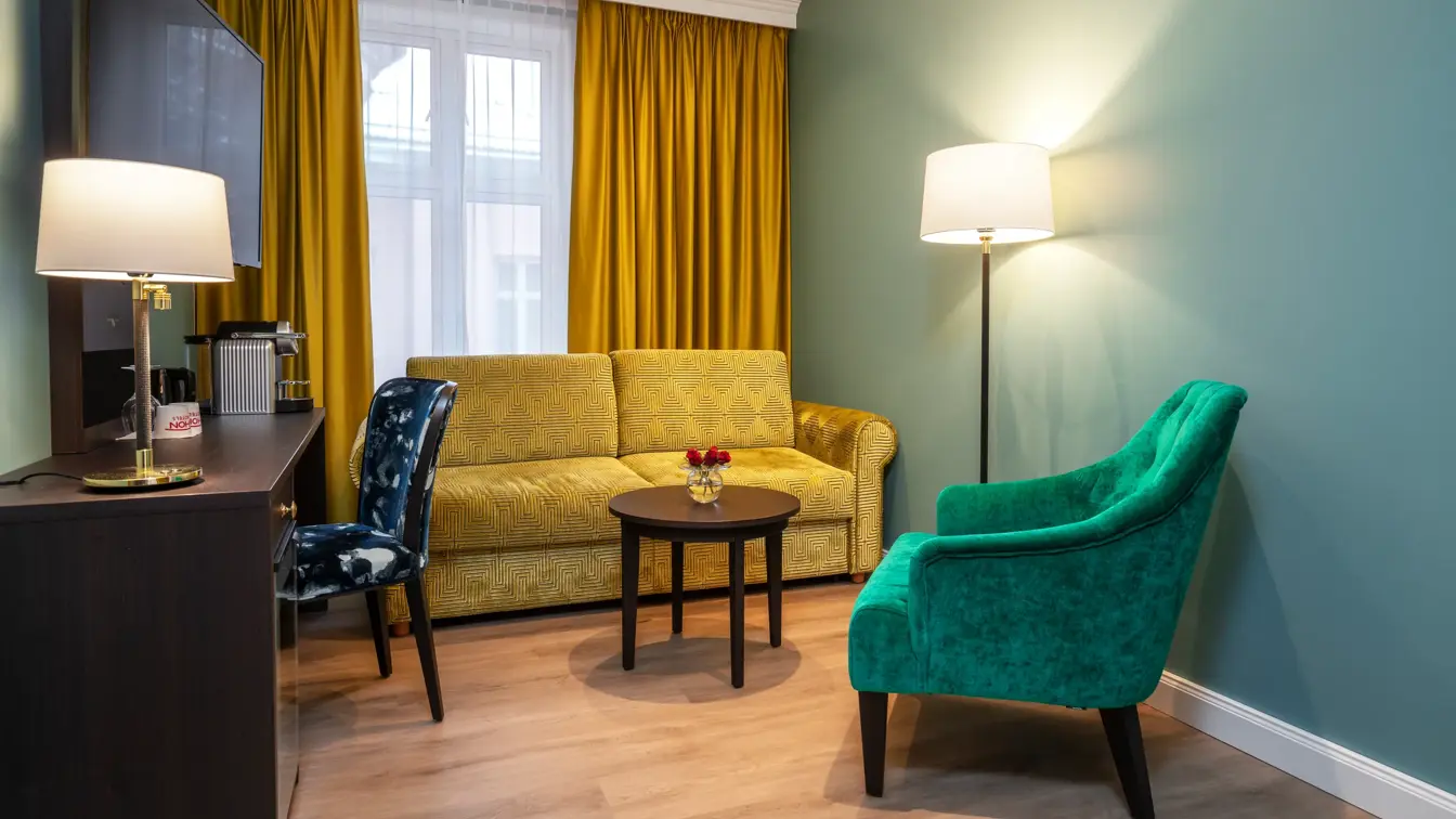 Stue med stort vindu, gule gardiner fra tak til gulv. Limegrønn sofa, rundt mørkt bord med røda blomster i vase. Grønn lenestol. Mørkt skrivebord med kaffemaskin, veggehengt TV over og mønstrete stol. I business rom på Thon Hotel Høyers i Skien. 