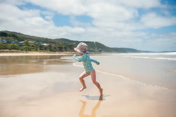 Small girl running up the sand from waves at the seaside. She is wearing a sun hat and long sleeves to protect her from the sun.