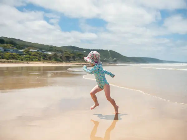 Small girl running up the sand from waves at the seaside. She is wearing a sun hat and long sleeves to protect her from the sun.
