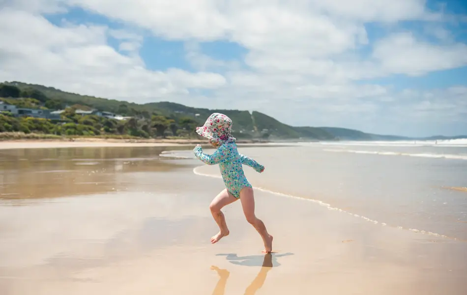 Small girl running up the sand from waves at the seaside. She is wearing a sun hat and long sleeves to protect her from the sun.