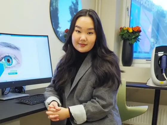 smilende dame som sitter i dressjakke foran en pc skjerm med bilde at et øye