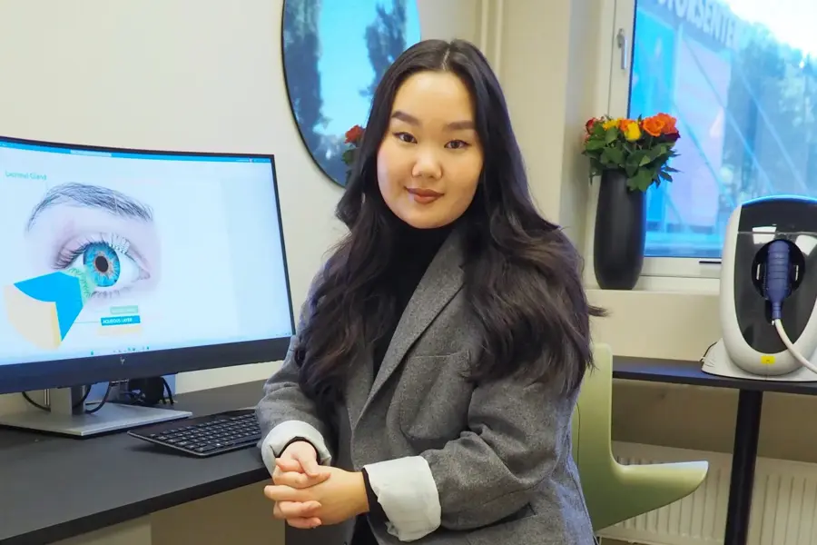 smilende dame som sitter i dressjakke foran en pc skjerm med bilde at et øye