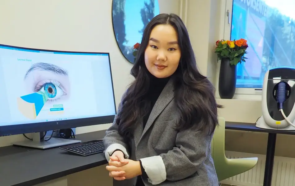 smilende dame som sitter i dressjakke foran en pc skjerm med bilde at et øye