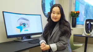 smilende dame som sitter i dressjakke foran en pc skjerm med bilde at et øye