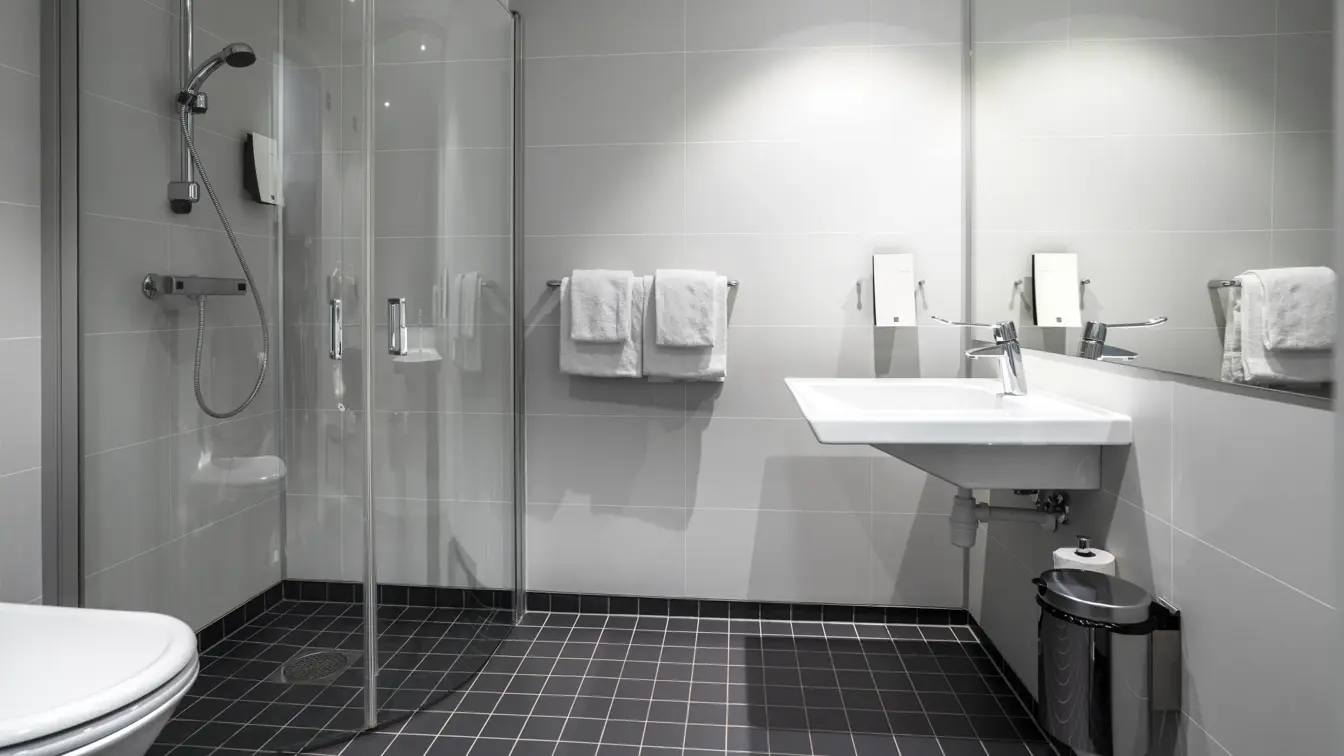 Bad på familierom med servant, stort speil, dusjhjørne og flislagt gulv.