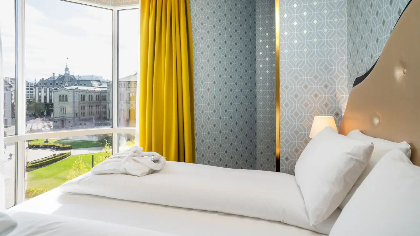 Dobbeltseng, mønsteret sølv tapet, gule gardiner og store vinduer med utsikt over Stortinget i junior suite på Thon Hotel Cecil