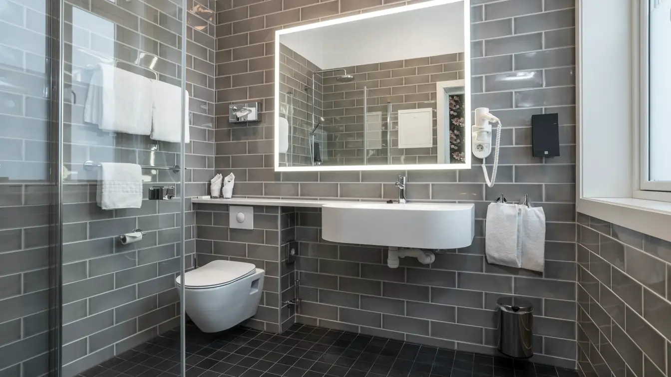Vegger og gulv med grå fliser, dusjhjørne med glassvegg, speil med LED lys rundt, hvit servant og vegghengt toalett. I standard baderom på Thon Hotel Høyers i Skien.