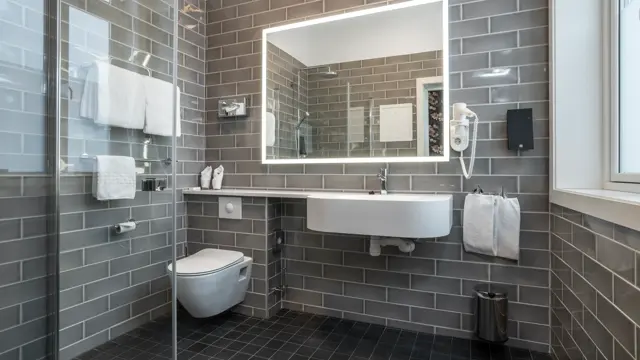 Vegger og gulv med grå fliser, dusjhjørne med glassvegg, speil med LED lys rundt, hvit servant og vegghengt toalett. I standard baderom på Thon Hotel Høyers i Skien.