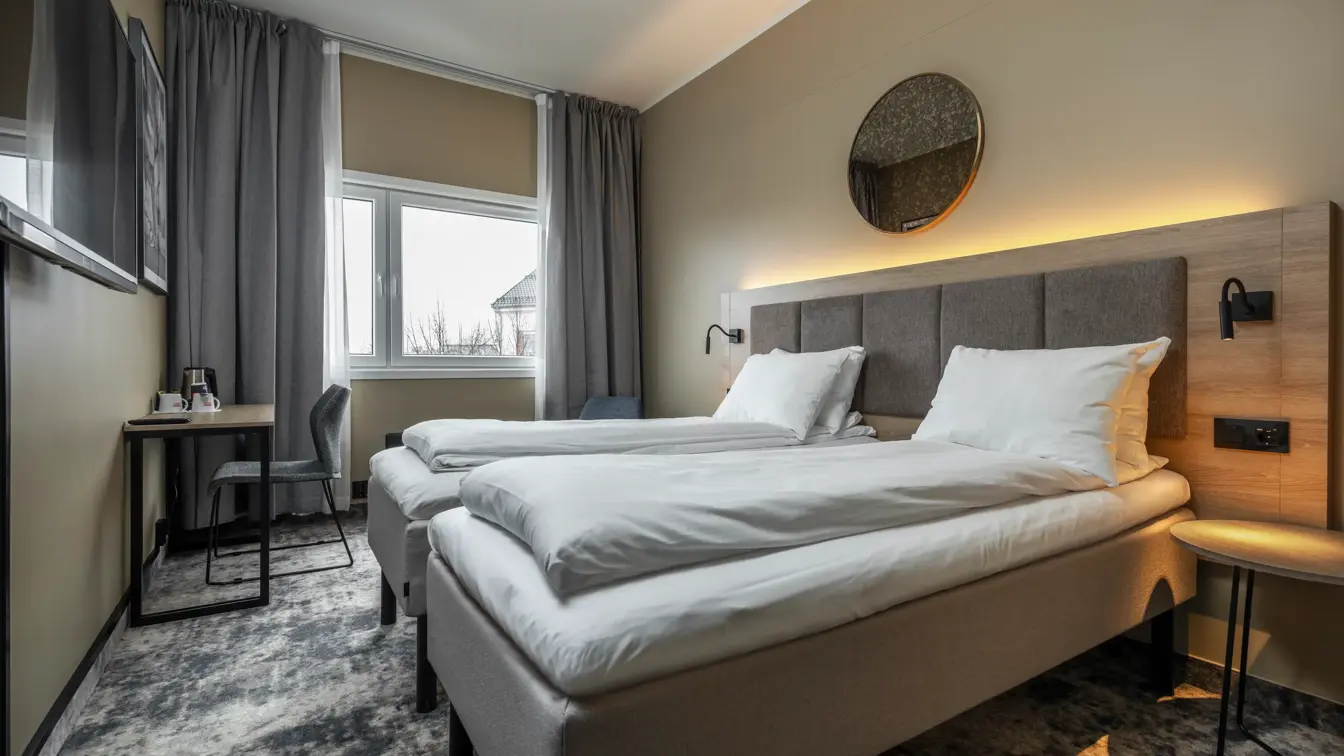 Dobbeltrom med twin senger, hvitt sengetøy, gulvteppe, lange gardiner, stort vindu og vegghengt TV.