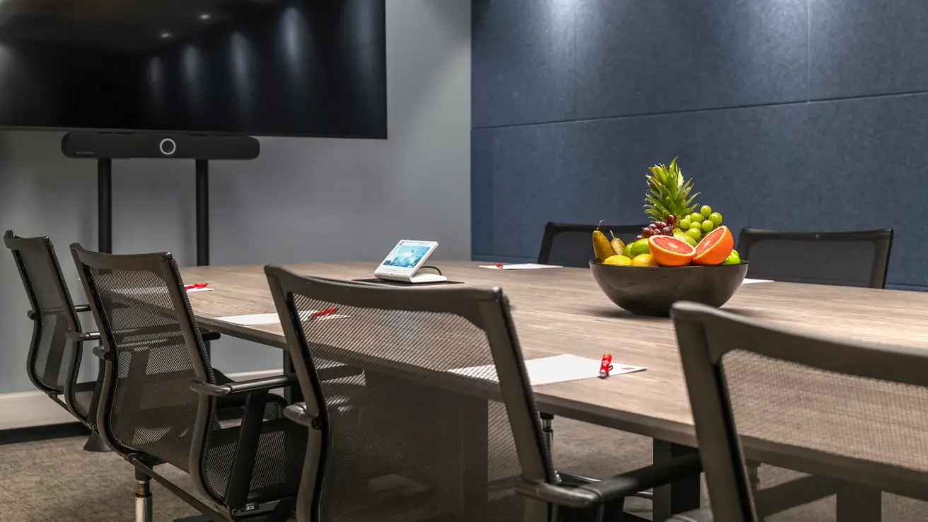Nærbilde av møterom med langbord, kontorstoler, bolle med frukt på bordet, blå vegg og stor TV skjerm.