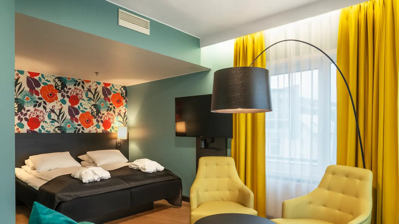 Blomstrende veggtapet, blå vegger, gule gardiner, to gule stoler, svart stuebord, stor svart lampe og stort glassvindu.