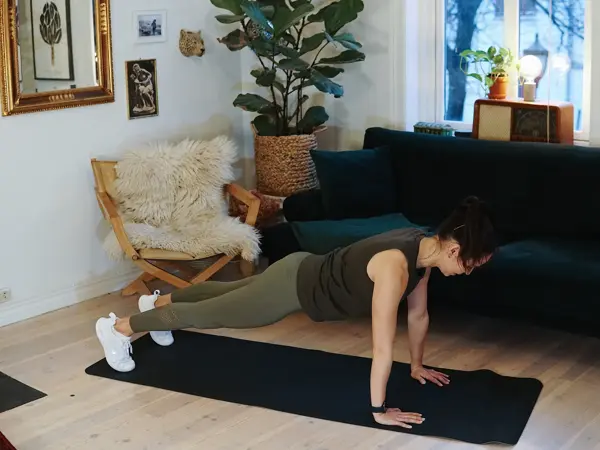 treningsekspert Anna Johansen trener på matte på gulvet, mørkegrønn sofa i bakgrunnen, lys stol med pels oppi, str grønn plante, bilder på veggen