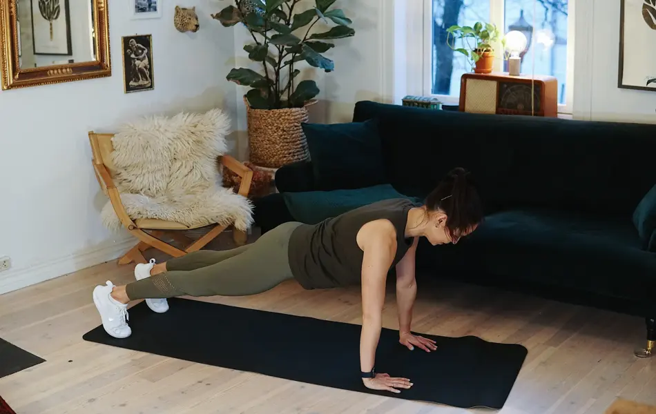 treningsekspert Anna Johansen trener på matte på gulvet, mørkegrønn sofa i bakgrunnen, lys stol med pels oppi, str grønn plante, bilder på veggen