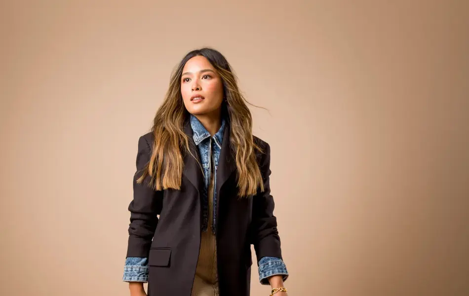 Tsustumi Hoang i mørk blå dressjakke, olaskjorte, beige skjørt, sort liten veske og brune semskede høye støvletter