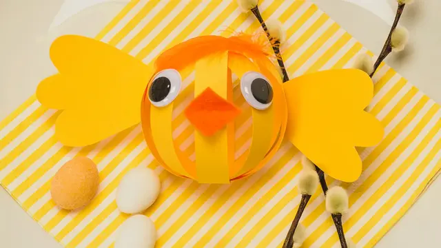 Papir-påskekylling laget av gult papir med oransje nebb og fjær til hår på en tallerken