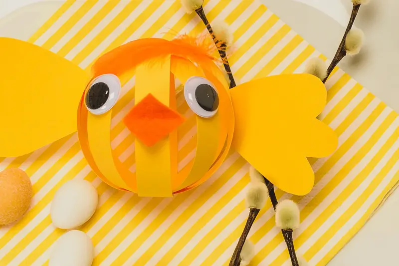Papir-påskekylling laget av gult papir med oransje nebb og fjær til hår på en tallerken