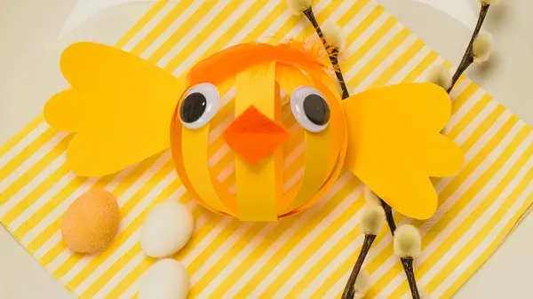 Papir-påskekylling laget av gult papir med oransje nebb og fjær til hår på en tallerken