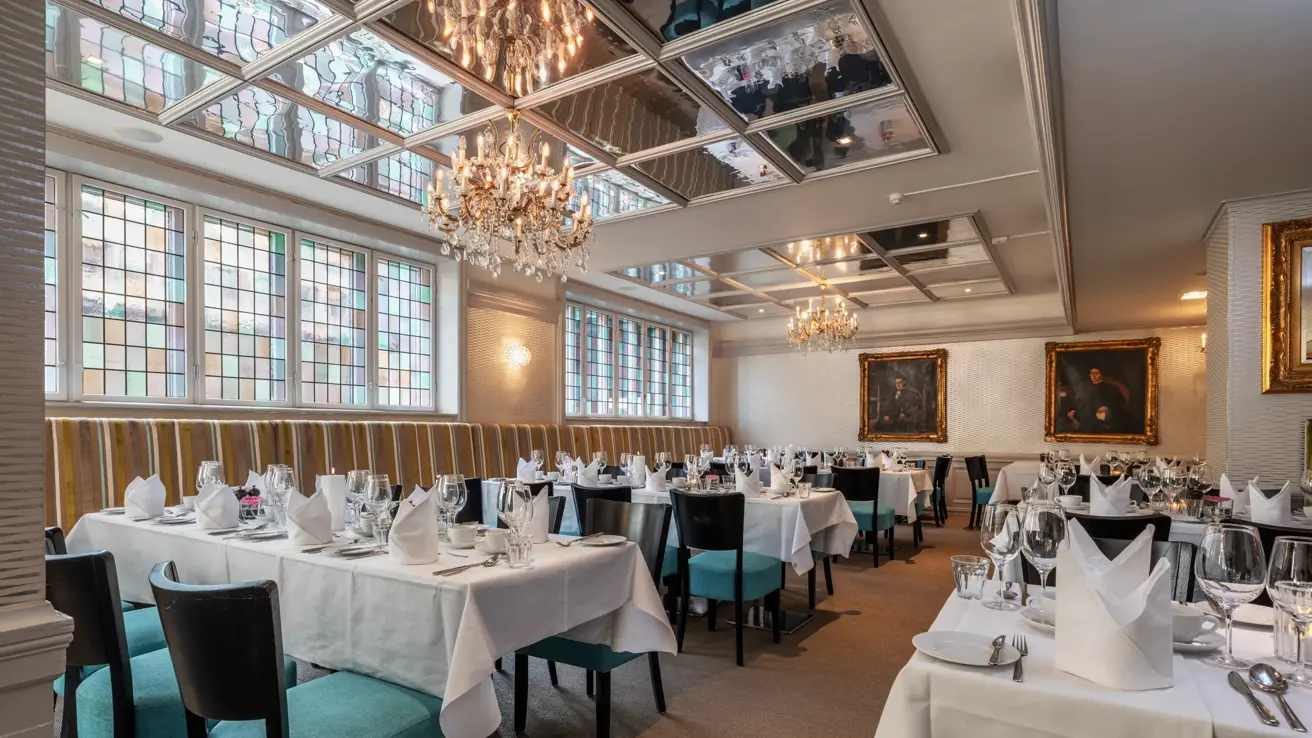 Restaurant dekket til selskap med hvite duker og turkise stoler. Lysekroner og speil i taket.