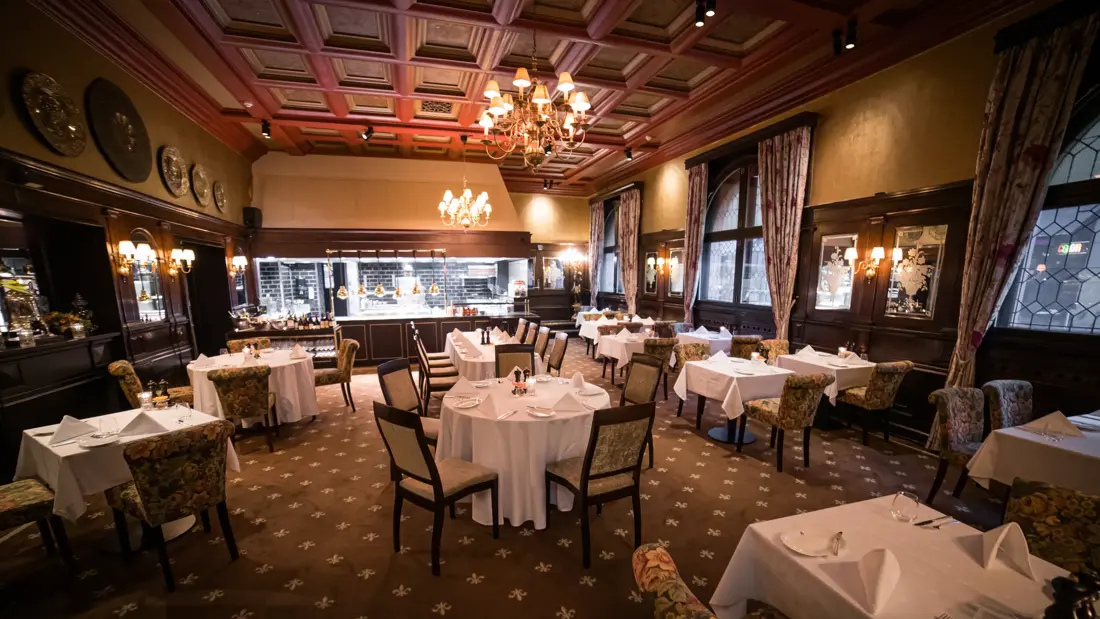 Restaurant med teppegulv, bord med hvite duker, brunt tak og lysekroner