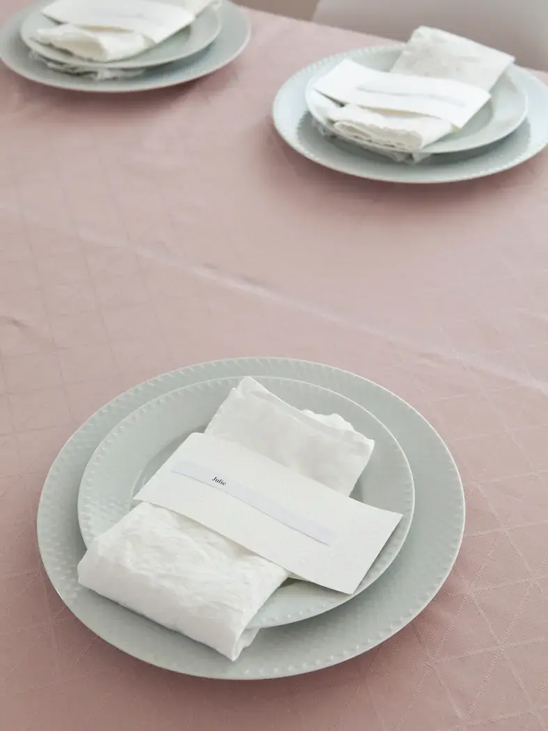 Steg 3 - Bord dekket med tallerkener og hvit linservietter og navnelapp på en rosa duk 