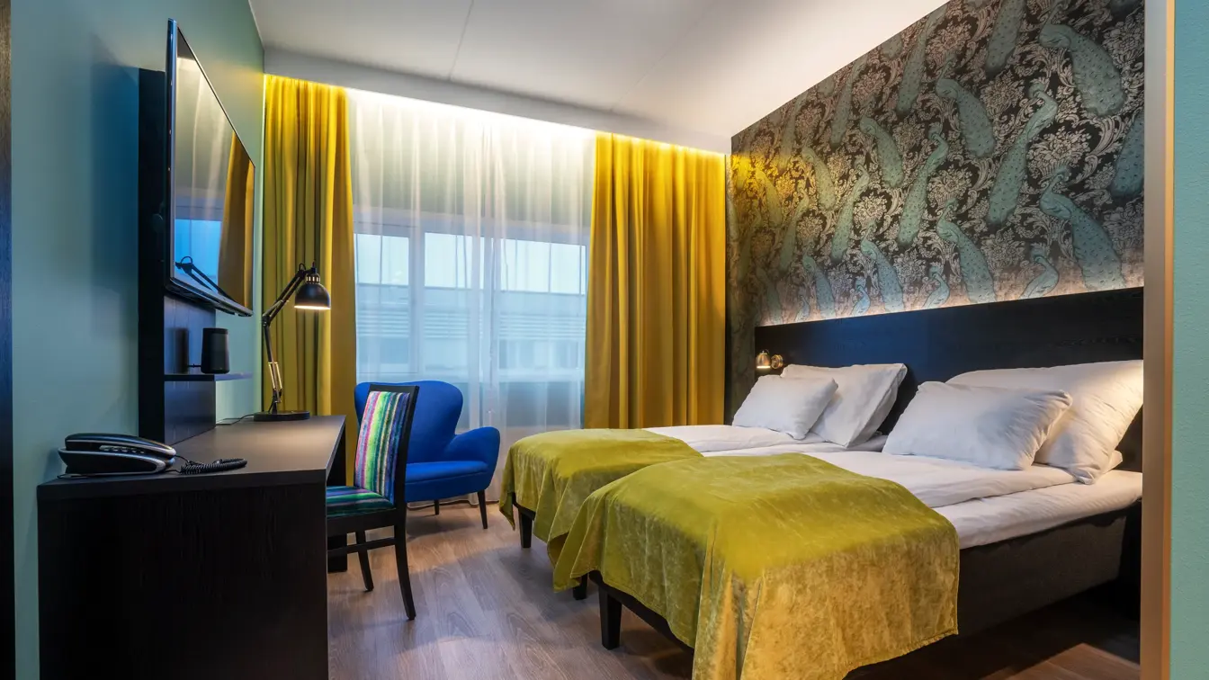 Parkettgulv, vindu fra gulv til tak, gule gardiner, veggspeil, to enkle senger, tapetvegg med mønster, skrivebord, to stoler. 