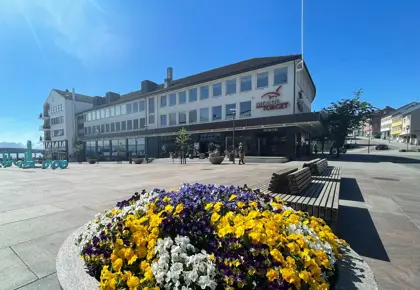 Fasadebilde av kjøpesenteret MoldeTorget. Bildet viser kjøpesenteret i bakgrunnen med sjø, blomster og kunst i forgrunnen.