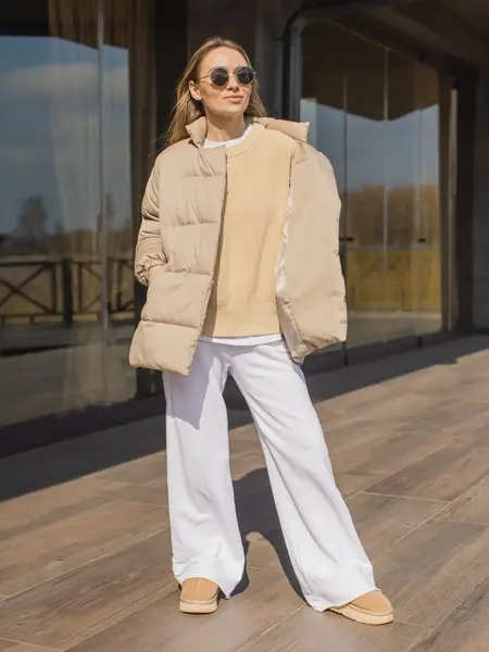 En dame i beige boblejakke og hvite bukser står på en solfylt terrasse