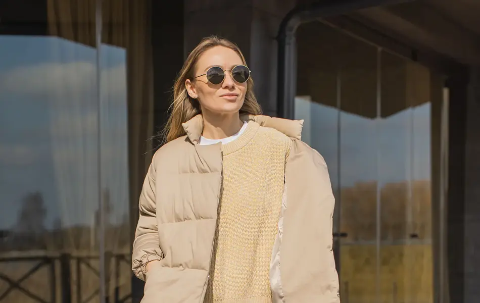 En dame i beige boblejakke og hvite bukser står på en solfylt terrasse