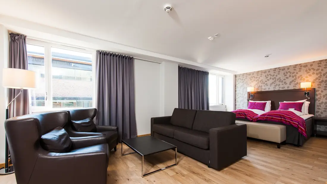 Dobbeltseng, tapetvegg, hvote vegger, vindu, grå gardiner, brun sofa, to brune lenestoler, salongbord