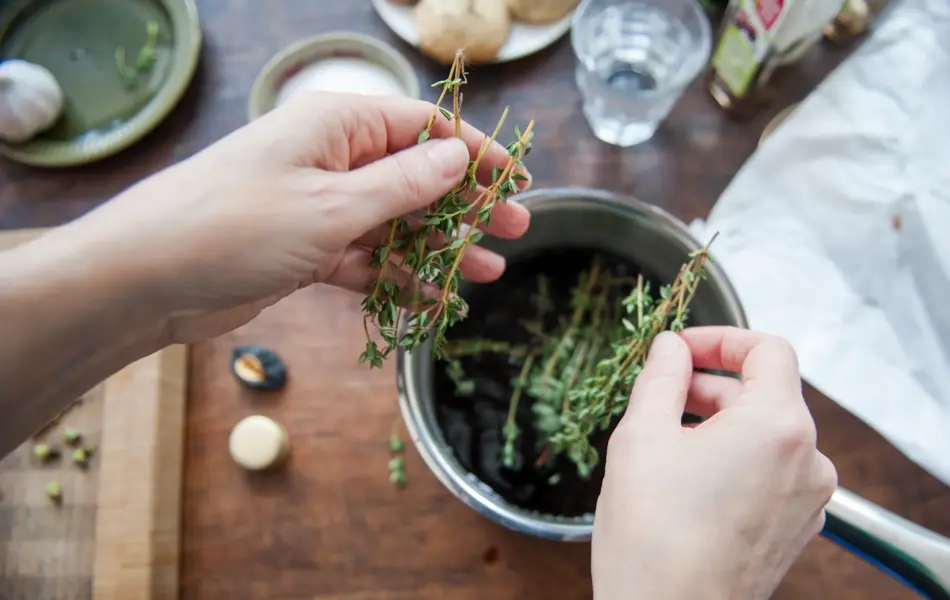 Woman adding fresh thyme leaves to a cooking pot filled with balsamic vinegar, to make a dressing. First person perspective.