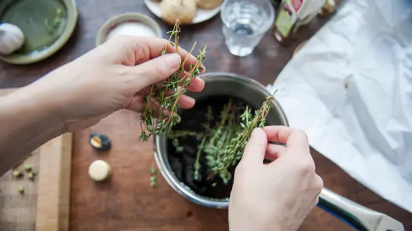 Woman adding fresh thyme leaves to a cooking pot filled with balsamic vinegar, to make a dressing. First person perspective.