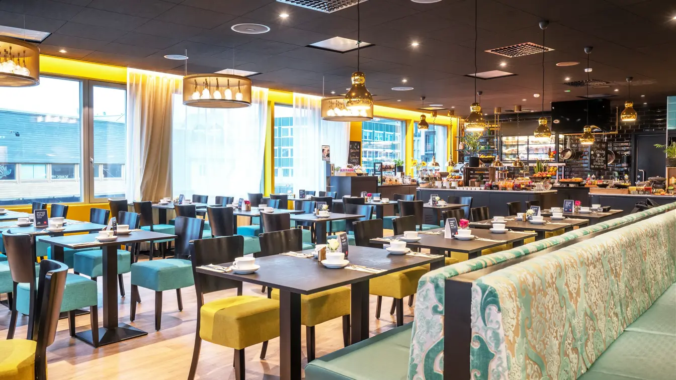 Restaurant med svarte spisebord, blå og gule stoler. vinduer fra gult til tak og store gule taklamper