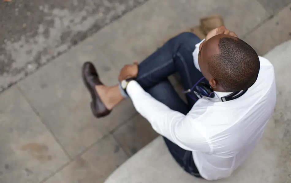 Mann med hvit skjorte, jeans og brune skinnsko sitter på stein sett ovenfra 