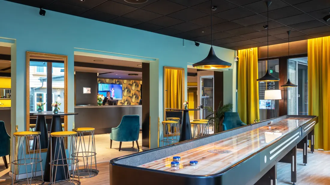 Shuffleboard, høge bord med gule barkrakker og turkise vegger i lobbyen på Thon Hotel Maritim