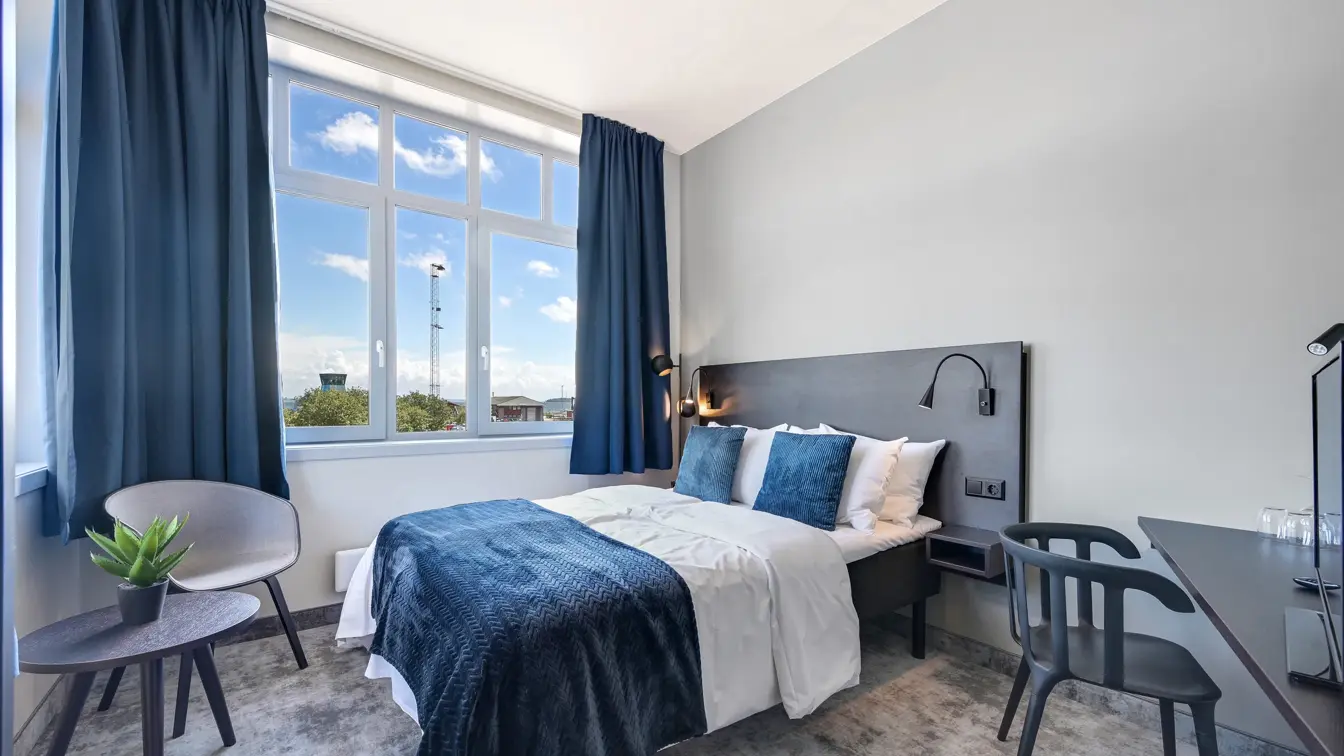 Dobbeltseng, hvite vegger, skrivebord, stol, TV,  vindu, blå gardiner