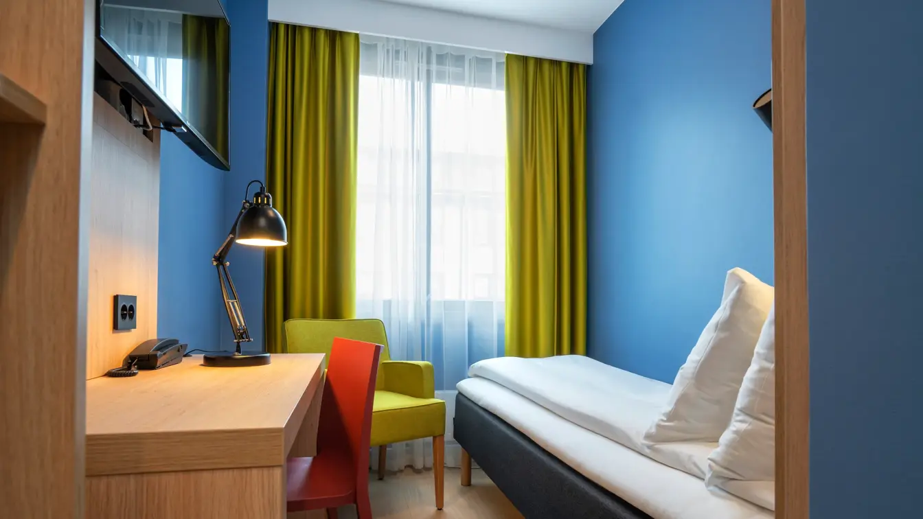 Garderobe, skrivebord, TV, blå vegger, gul stol, gule gardiner og enkeltseng på lite enkeltrom på Thon Hotel Astoria