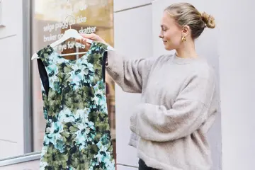 Dame holder opp en kjole med grønt blomstermønster utenfor en bruktbutikk