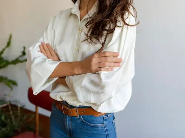 Nærbilde av overkroppen til en kvinne. Hun har armene i kors og brunt hår som henger løst ned over skuldrene. Hun er iført en hvit bluse, jeans og et brunt skinnbelte.
