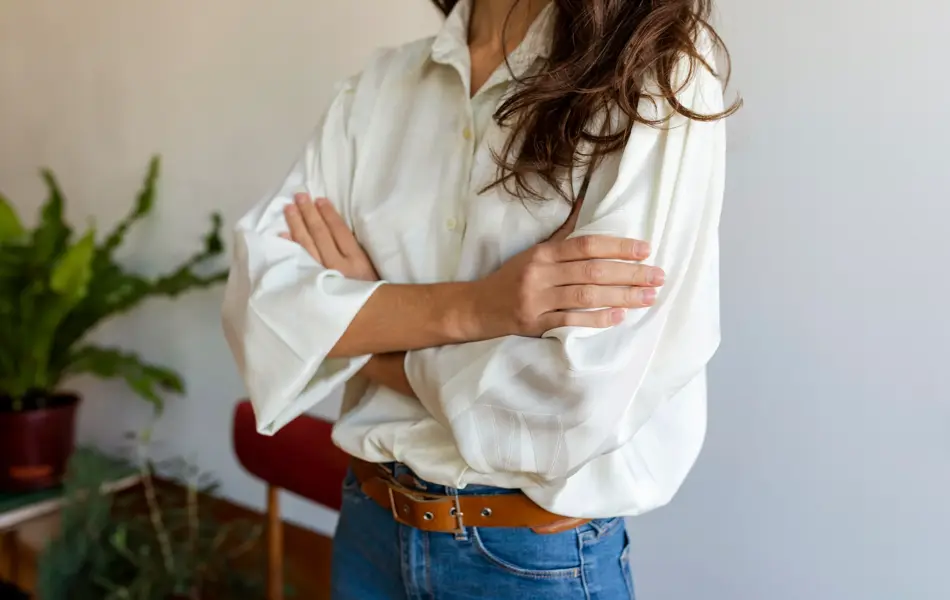 Nærbilde av overkroppen til en kvinne. Hun har armene i kors og brunt hår som henger løst ned over skuldrene. Hun er iført en hvit bluse, jeans og et brunt skinnbelte.