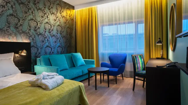 Parkettgulv, turkis sofa, to stoler, salongbord, skrivebord, dobbeltseng, vindu fra gulv til tak, gule gardiner, tapetvegg med mønster 