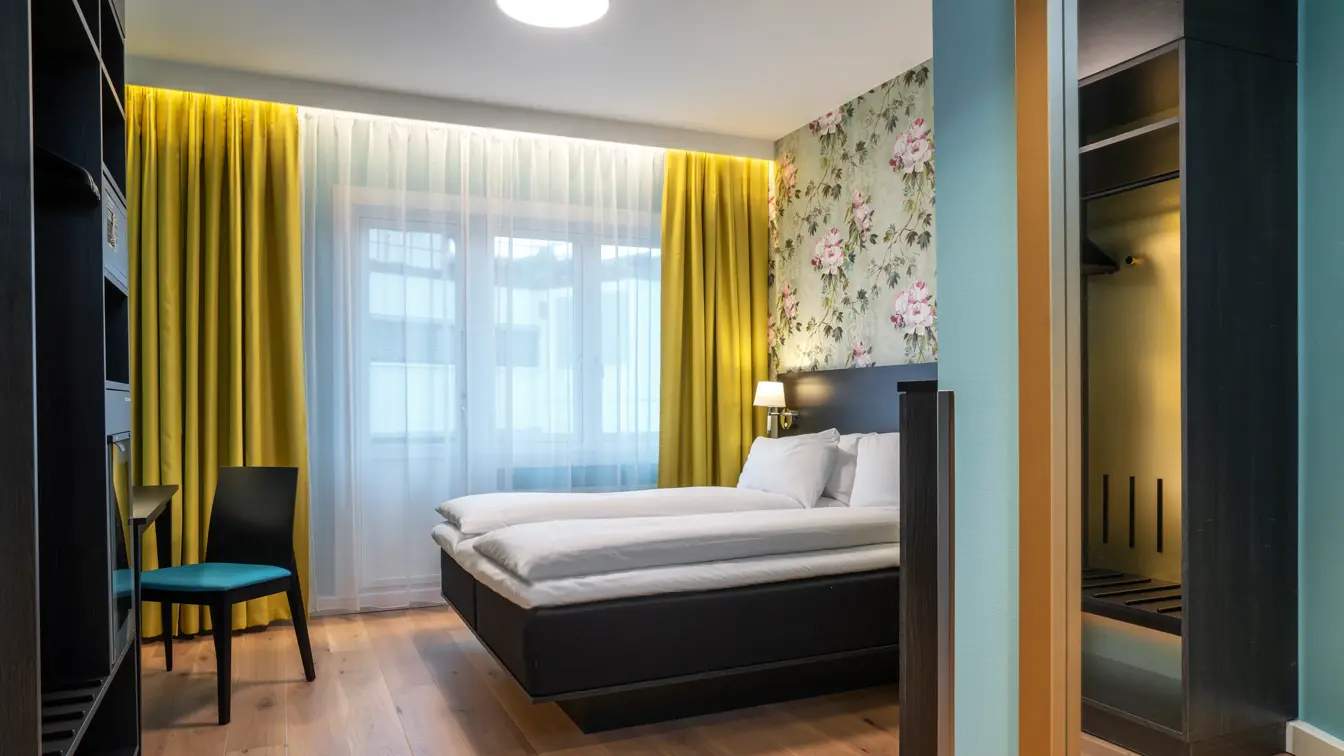 Vindu fra gulv til tak, gule gardiner, dobbeltseng, skrivebord, stol, speil på vegg, blomstrete tapetvegg, parkettgulv. 