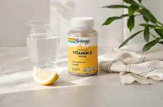 En boks med Vitamin C fra Solaray står på en benk med et glass vann og en skive sitron.