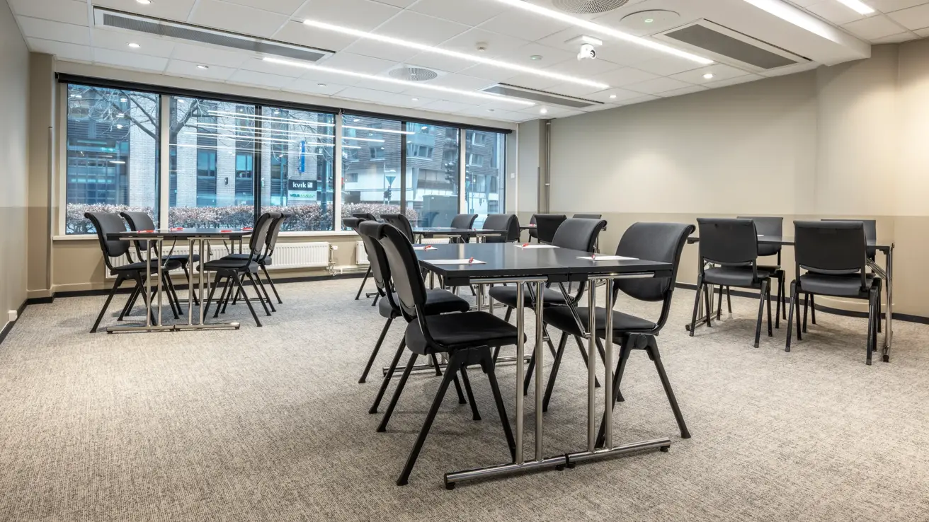 Møterom med flere gruppebord, vinduer langs hele veggen i enden med utsikt til gaten utenfor.