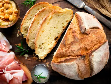 Rustic loaf of homemade bread served with olive oil, rosemary and cured ham on dark wooden table. Overhead view