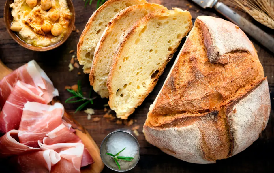 Rustic loaf of homemade bread served with olive oil, rosemary and cured ham on dark wooden table. Overhead view