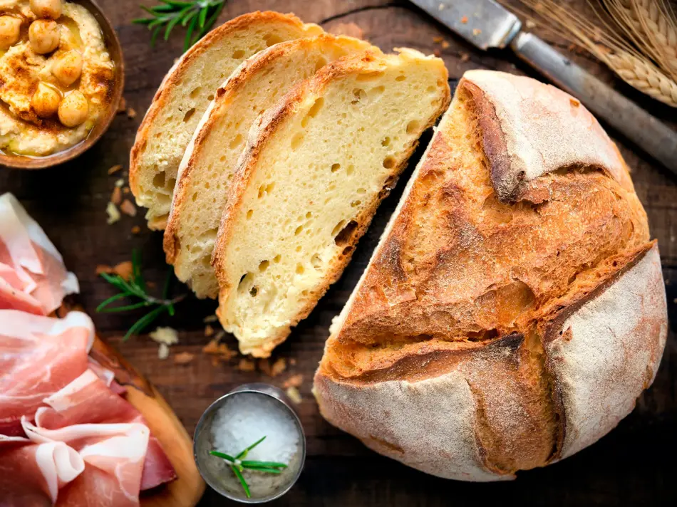 Rustic loaf of homemade bread served with olive oil, rosemary and cured ham on dark wooden table. Overhead view