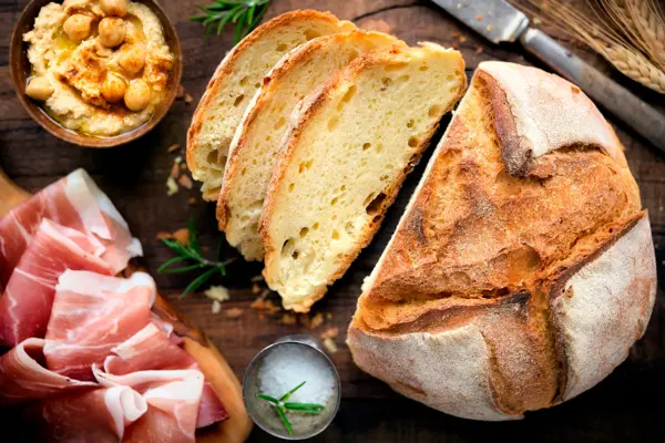 Rustic loaf of homemade bread served with olive oil, rosemary and cured ham on dark wooden table. Overhead view