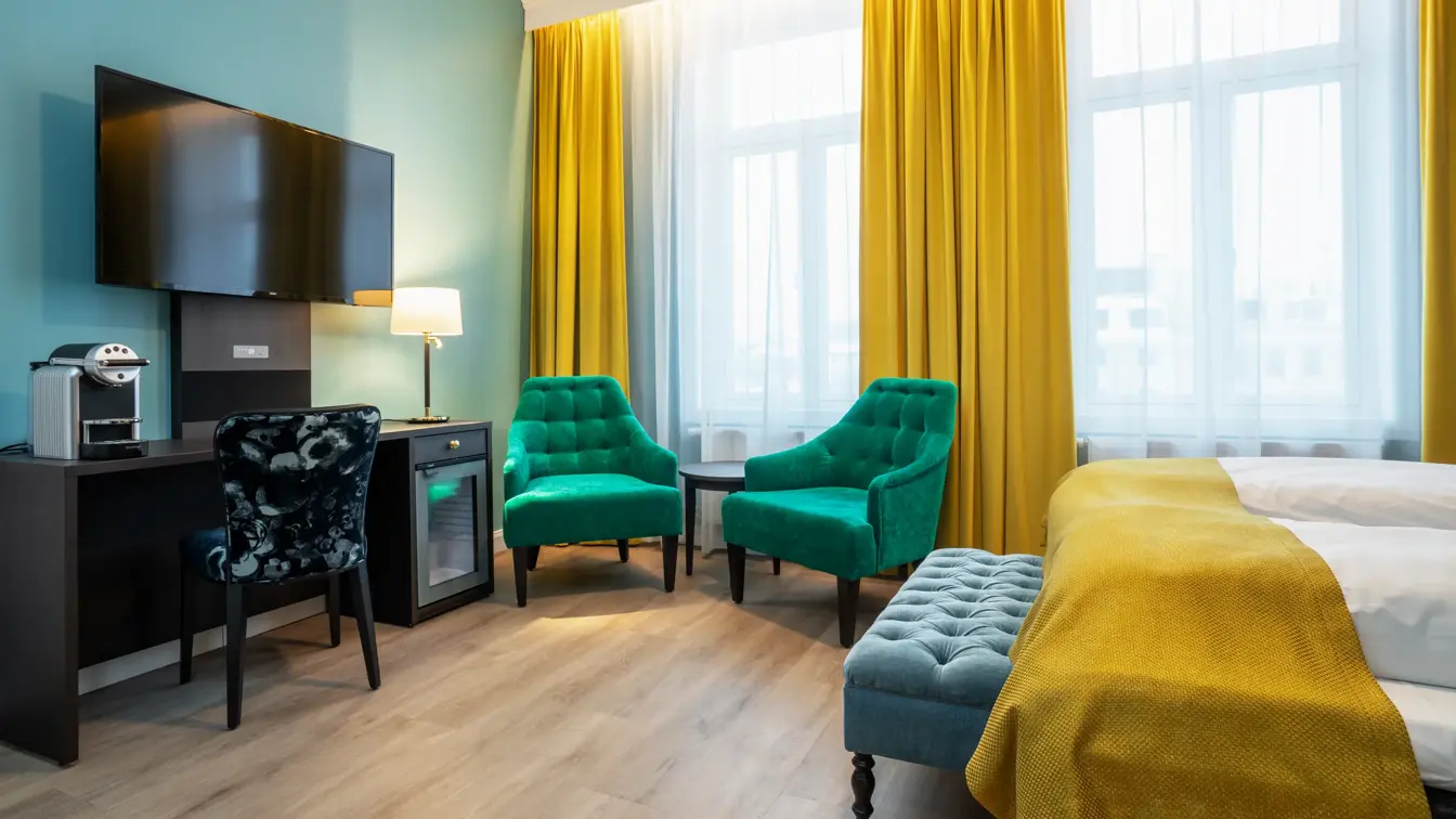 Store vinduer med gule gardiner fra tak til gulv og to grønne lenestoler. Mørkt skrivebord med kaffemaskin, minibar, veggehengt TV over og mørk mønstret stol. Dobbeltseng med gult sengeteppe. I superior rom på Thon Hotel Høyers i Skien.