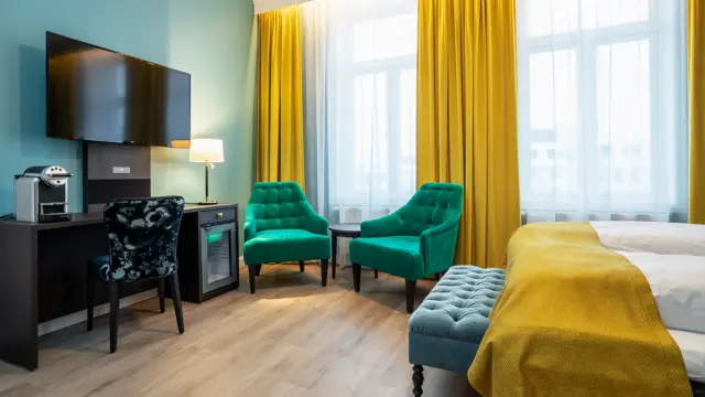Store vinduer med gule gardiner fra tak til gulv og to grønne lenestoler. Mørkt skrivebord med kaffemaskin, minibar, veggehengt TV over og mørk mønstret stol. Dobbeltseng med gult sengeteppe. I superior rom på Thon Hotel Høyers i Skien.